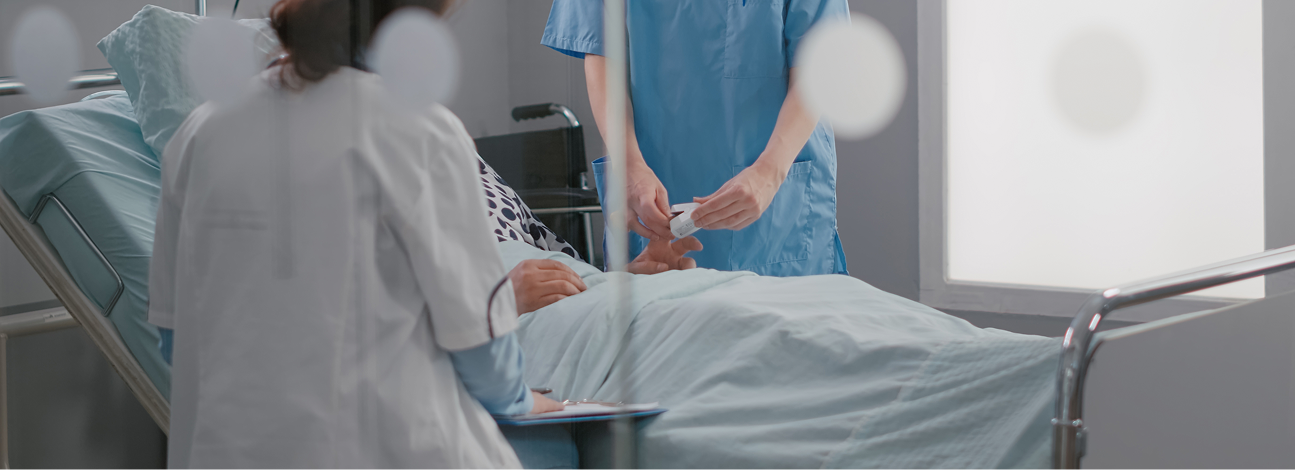 Patient lies in a hospital bed while healthcare staff in blue scrubs provide care at the bedside. One staff member stands near the patient’s arm holding a small medical item, while another sits beside the bed writing on a clipboard. The scene is viewed through a glass partition with soft circular reflections, in a bright room with a window and clinical equipment nearby.